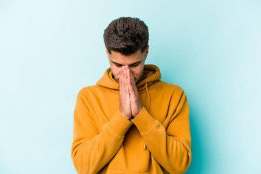 Kafkasyalı genç adam mavi arka planda izole edilmiş dua ediyor, bağlılık gösteriyor, dindar biri ilahi ilham arıyor..