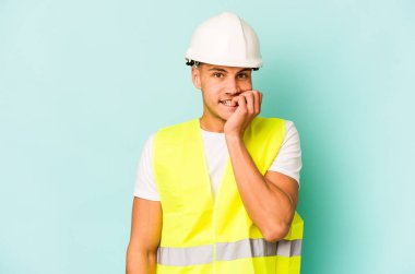 Kafkasyalı genç işçi, mavi arka planda izole edilmiş tırnak yiyor, gergin ve endişeli..