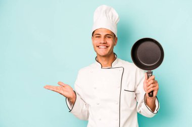 Kafkasyalı genç aşçı elinde uçan tavayla mavi arka planda izole edilmiş bir şekilde avuçta fotokopi alanı ve belinde bir el daha gösteriyor..