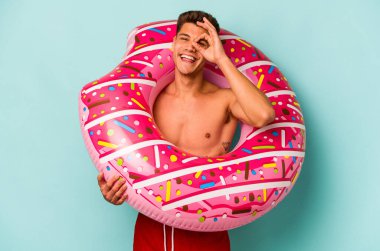 Mavi arka planda izole edilmiş şişirilebilir donut tutan genç beyaz bir adam heyecanlı..