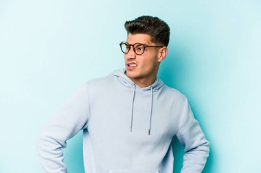 Kafkasyalı genç adam mavi arka planda izole, kafası karışmış, şüpheli ve güvensiz hissediyor..
