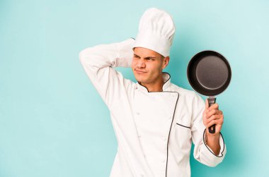 Kafkasyalı genç aşçı elinde uçan tavayla mavi arka planda izole edilmiş kafasının arkasına dokunuyor, düşünüyor ve bir seçim yapıyor..