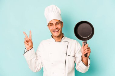 Kafkasyalı genç aşçı elinde uçan tavayla mavi arka planda izole edilmiş neşeli ve tasasız parmaklarıyla barış sembolü gösteriyor..