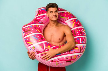 Kafkasyalı genç adam mavi arka planda izole edilmiş şişirilebilir bir çörek tutuyor. Amaçlarına ve amaçlarına ulaşma hayalleri kuruyor.