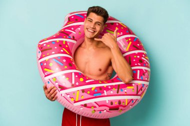 Mavi arka planda izole edilmiş şişme donut tutan genç beyaz bir adam parmaklarıyla bir cep telefonu hareketi gösteriyor..