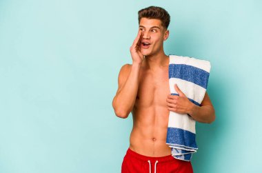 Mavi arka planda izole edilmiş plaj havlusu tutan genç beyaz adam gizli bir sıcak fren haberi veriyor ve yan bakıyor.