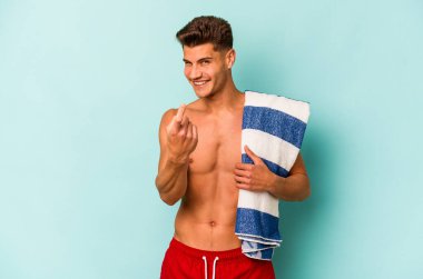 Elinde plaj havlusu tutan genç beyaz adam, mavi arka planda izole edilmiş, seni işaret ediyor, sanki seni davet ediyormuş gibi..