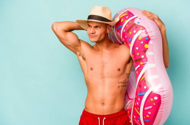 Kafkasyalı genç bir adam mavi arka planda izole edilmiş şişirilebilir bir çörek tutuyor. Kafasının arkasına dokunuyor, düşünüyor ve bir seçim yapıyor..