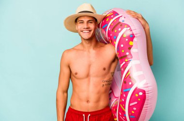 Mavi arka planda izole edilmiş şişme bir çörek tutan genç beyaz adam mutlu, güler yüzlü ve neşeli..