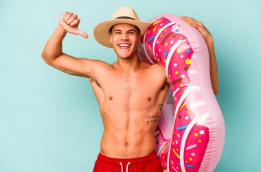 Mavi arka planda izole edilmiş şişme bir çörek tutan genç beyaz adam gururlu ve kendinden emin hissediyor, örnek olarak.