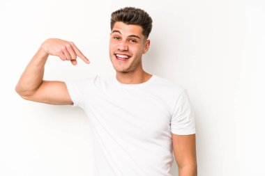 Beyaz arka planda yaşayan beyaz tenli genç bir adam gömlek fotokopi alanını gösteriyor, gururlu ve kendinden emin.