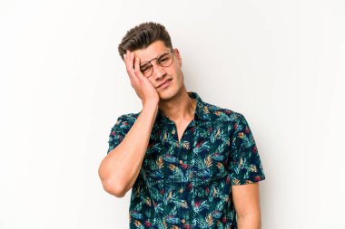 Beyaz arka planda izole edilmiş genç beyaz bir adam yorgun ve uykulu bir şekilde elini başının üstünde tutuyor..