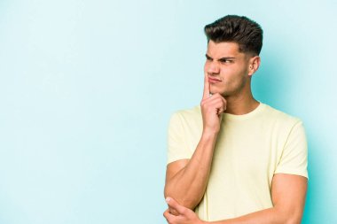 Mavi arka planda izole edilmiş genç beyaz adam şüpheli ve şüpheci bir ifadeyle yana bakıyor..