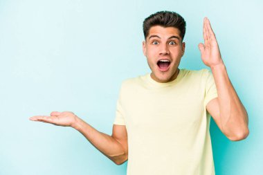 Mavi arka planda izole edilmiş genç beyaz bir adam avucunun içinde fotokopi alanı tutuyor, el üstünde tutuyor. Şaşırmış ve mutlu..