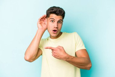 Mavi arka planda izole edilmiş genç beyaz adam bir dedikoduyu dinlemeye çalışıyor..