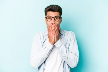 Mavi arka planda izole edilmiş genç beyaz adam elleriyle ağzını kapatırken şok oldu..