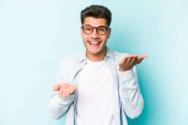 Mavi arka planda izole genç beyaz adam kollarıyla ölçek yapar, mutlu ve kendinden emin hisseder..