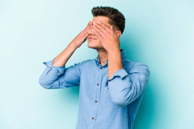 Mavi arka planda izole edilmiş genç beyaz adam elleriyle gözlerini kapatmaktan korkuyor..