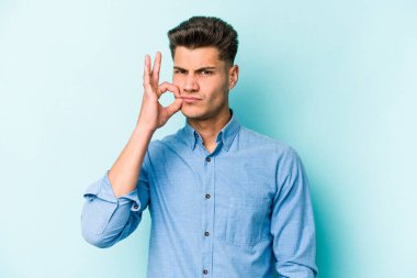 Genç beyaz adam mavi arka planda izole edilmiş ve parmakları dudaklarında bir sır saklıyor..