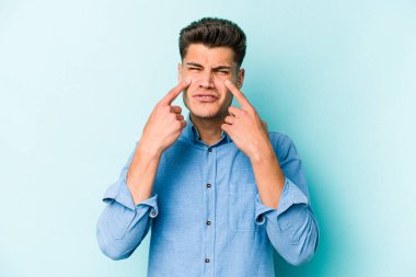 Kafkasyalı genç adam mavi arka planda izole edilmiş, ağlıyor, bir şeyden memnun değil, acı ve kafa karışıklığı konsepti..