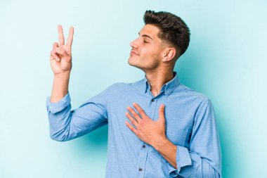 Kafkasyalı genç adam mavi arka planda izole edilmiş yemin ediyor, elini göğsüne koyuyor..
