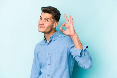 Kafkasyalı genç bir adam mavi arka planda izole edilmiş bir şekilde göz açıp kapayana kadar iyi bir el hareketi yapıyor..