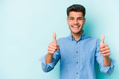 Mavi arka planda izole edilmiş genç beyaz adam gülümsüyor ve başparmağını kaldırıyor.