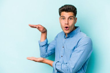 Mavi arka planda izole genç beyaz adam şok ve hayret içinde elleri arasında bir kopya boşluk tutuyor.