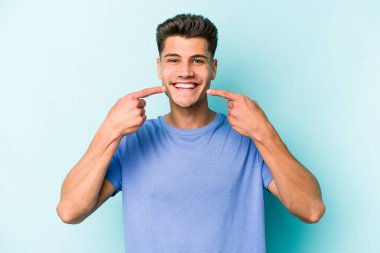 Mavi arka planda izole edilmiş genç beyaz adam gülümsüyor, parmaklarını ağıza doğrultuyor..