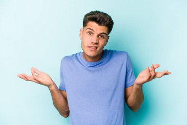 Kafkasyalı genç adam mavi arka planda izole edilmiş sorgulama jestinde şüphe ediyor ve omuz silkiyor..