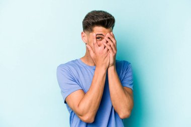 Mavi arka planda izole edilmiş genç beyaz adam kameraya parmaklarıyla göz kırpıyor, yüzünü kapatmaktan utanıyor..