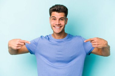 Mavi arka planda izole edilmiş genç beyaz adam bir gömlek fotokopi alanını işaret ediyor, gururlu ve kendinden emin.