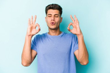 Mavi arka planda izole edilmiş genç beyaz adam yorucu bir günün ardından rahatlıyor. Yoga yapıyor..