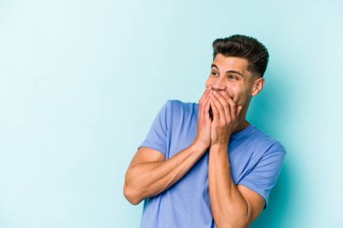 Mavi arka planda izole edilmiş genç beyaz bir adam bir şeye gülüyor, ağızlarını ellerle kapatıyor..