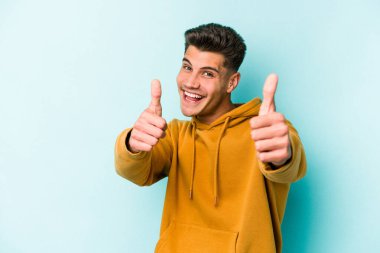 Kafkasyalı genç adam mavi arka planda izole edilmiş. Baş parmağını kaldırıyor, gülümsüyor ve kendinden emin..