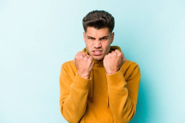 Kafkasyalı genç bir adam mavi arka planda izole edilmiş. Yumruk atıyor, sinirleniyor, kavga ediyor, kavga ediyor..