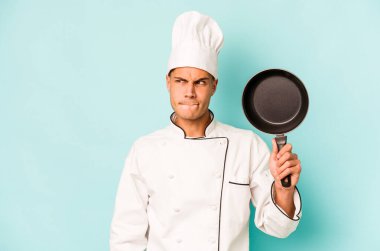 Kafkasyalı genç aşçı elinde uçan tavayla mavi arka planda izole edilmiş, kafası karışmış, şüpheli ve güvensiz hissediyor..