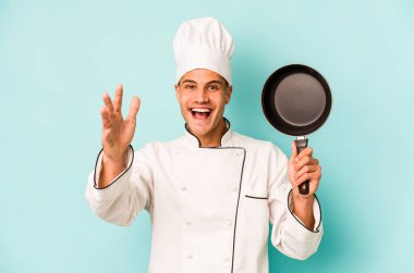 Kafkasyalı genç aşçı elinde uçan bir tavayla mavi arka planda izole edilmiş güzel bir sürpriz bekliyor, heyecanlı ve el kaldırıyor..