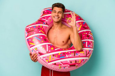 Mavi arka planda izole edilmiş şişme donut tutan beyaz bir genç adam neşeli ve kendinden emin..