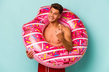 Mavi arka planda izole edilmiş şişme donut tutan genç beyaz bir adam sanki seni davet ediyormuş gibi seni işaret ediyor..