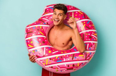 Mavi arka planda izole edilmiş şişme donut tutan genç beyaz bir adam bir fikri, ilham verici bir konsepti var..