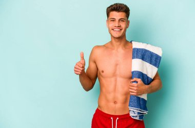 Elinde plaj havlusu tutan genç beyaz adam mavi arka planda izole edilmiş gülümsüyor ve başparmağını kaldırıyor.