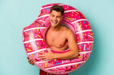 Mavi arka planda izole edilmiş şişme bir çörek tutan genç beyaz adam gülüyor ve eğleniyor..