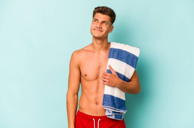 Elinde plaj havlusu tutan genç beyaz adam mavi arka planda izole edilmiş hedeflere ve amaçlara ulaşmayı hayal ediyor.