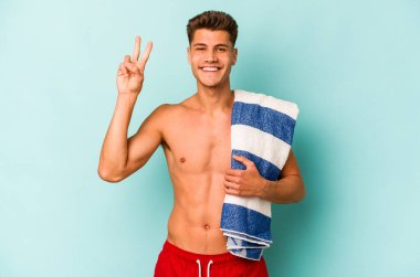 Elinde plaj havlusu tutan genç beyaz adam mavi arka planda iki numarayı parmaklarıyla gösteriyor..