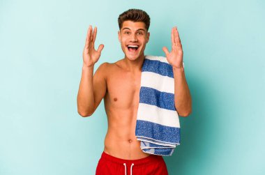 Mavi arka planda kumsal havlusu tutan genç beyaz adam hoş bir sürpriz alıyor, heyecanlı ve el kaldırıyor..
