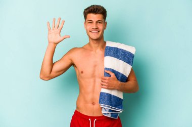 Elinde plaj havlusu tutan genç beyaz adam mavi arka planda izole edilmiş gülümseyen beş numarayı parmaklarıyla gösteriyor..