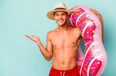 Mavi arka planda izole edilmiş şişme donut tutan genç beyaz bir adam bir avuç içinde fotokopi alanı ve belinde başka bir el tutuyor..