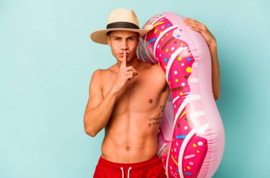 Mavi arka planda izole edilmiş şişme donut tutan genç beyaz bir adam bir sır saklıyor ya da sessizlik istiyor..