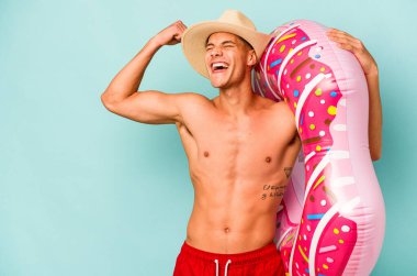 Mavi arka planda izole edilmiş şişirilebilir donut tutan genç beyaz adam zafer kazandıktan sonra yumruğunu kaldırıyor..